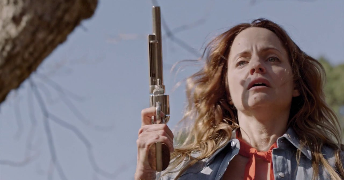 Mena Suvari as Cassandra, a griefing mother holding a revolver wearing a blue demin shirt witha an orange scarf.