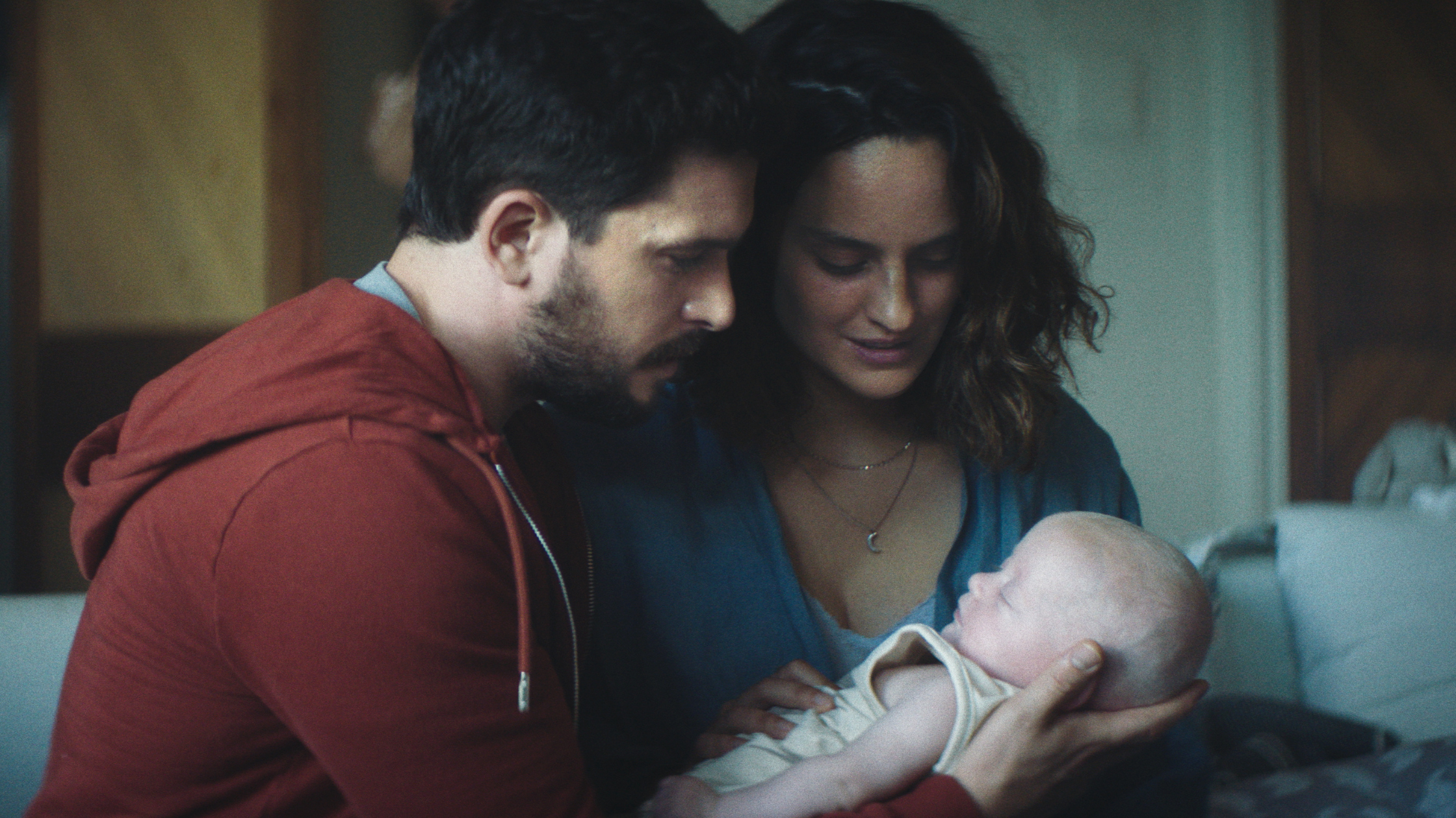 Josephine and Spencer welcome their new baby girl, Ruby, home from the hospital. Spencer is wearing a red hoodie sweater with a gray shirt, cradling Ruby in his arms, while Josphine smiles and coos at her.