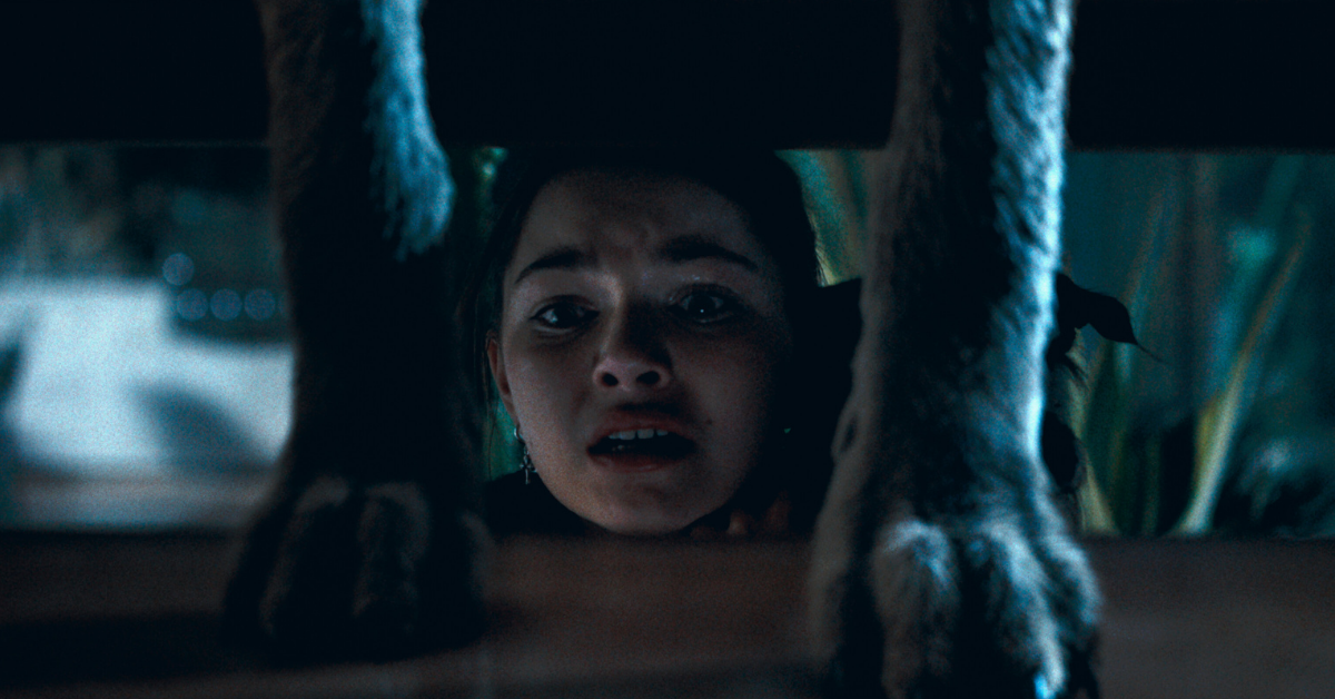 Mila Harris as Chloe in Coyotes (2025) hiding under the stairs with her dog as ravenous coyotes descend on her home.